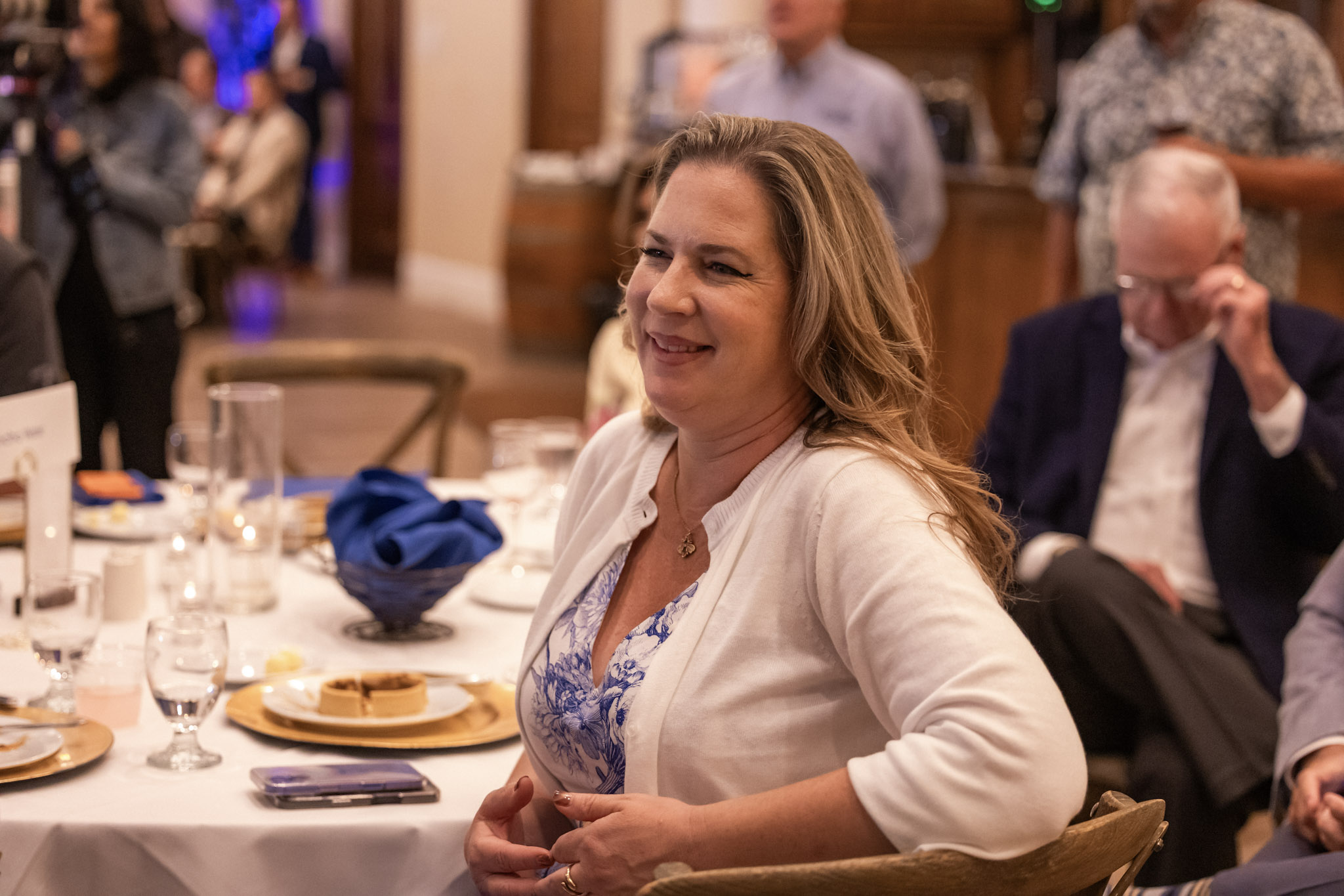SCWC-138 A woman in a white cardigan smiles while sitting at a recent luncheon